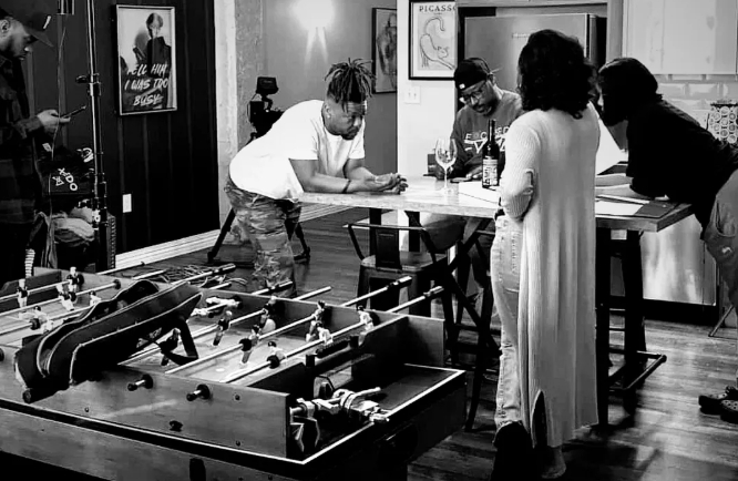 Black-and-white behind-the-scenes photo of the cast and camera crew gathered around a table