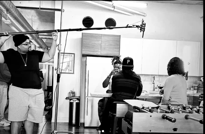 Black-and-white behind-the-scenes photo of a boom operator and actors filming in a kitchen